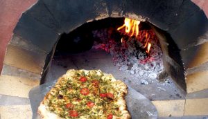 A pesto pizza being placed on an 800°F stone to cook. A\J AlternativesJournal.ca