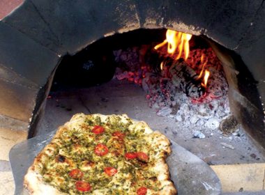 A pesto pizza being placed on an 800°F stone to cook. A\J AlternativesJournal.ca