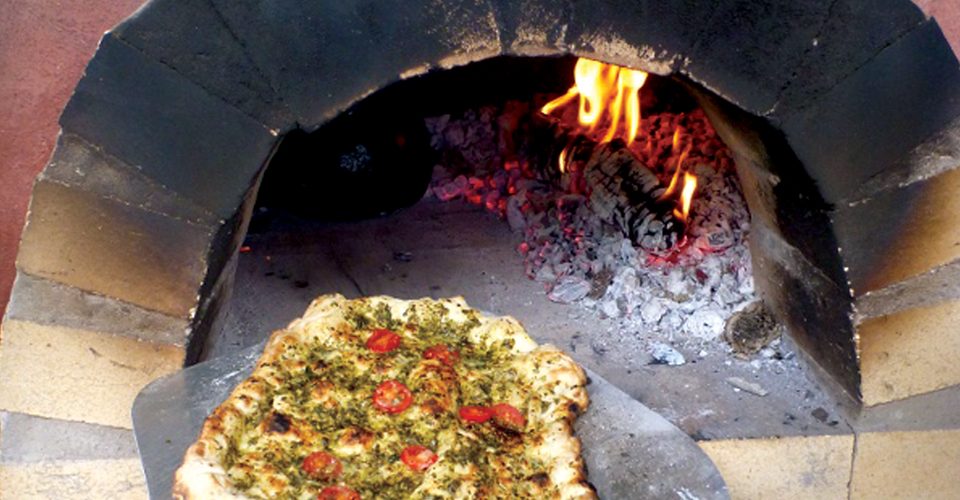 A pesto pizza being placed on an 800°F stone to cook. A\J AlternativesJournal.ca